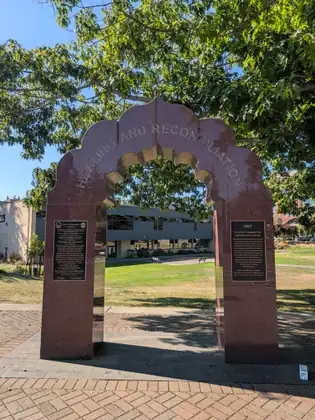 The red granite Healing and Reconciliation arch standing on a brick plaza, a plaque on each leg, under a big shade tree.