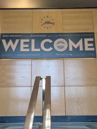 A long blue sign reading WELCOME, with the City of Bellingham seal as the O and the word for welcome in a dozen languages above and below it, under a clock on a wood-panelled wall.