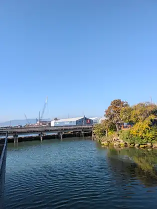 A calm stretch of harbour water under a clear blue sky, a low concrete bridge crossing it, with waterfront sheds, sailboat masts and a crane beyond and hills in the distance.