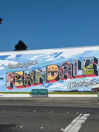 A mural the length of a building reading Greetings from Ferndale, Washington, each big letter filled with a local scene — flowers, a bird of prey, a bridge, berries, a totem pole and a cow — with a green bench in front of it.
