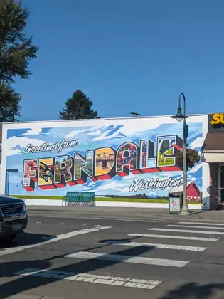 The Greetings from Ferndale, Washington mural seen whole from across the street, a green bench and a painted trash can below it, a lamp post hung with a flower basket, and a crosswalk and the front of a parked SUV in the foreground.