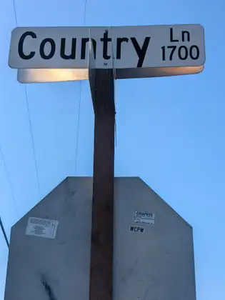 A street sign reading Country Ln 1700 on a wooden post, above the back of a stop sign, against a clear blue sky.