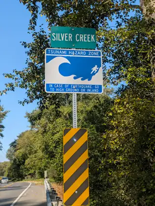 A green Silver Creek sign above a blue tsunami hazard zone sign and a yellow and black striped marker, at the edge of the road.