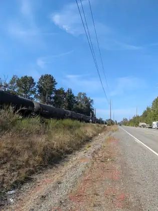 A long line of black tank cars on the tracks beside the road, the gravel shoulder running on ahead under power lines.