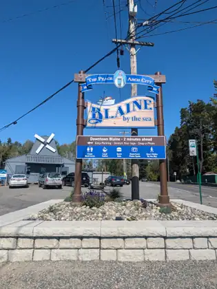 A tall roadside sign reading BLAINE by the sea, under a banner reading The Peace Arch City, with a panel below it reading Downtown Blaine, 2 minutes ahead; parked cars and a grey building stand behind.