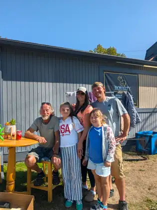 Five people smiling together at their yard sale: an older man sitting on a wooden stool, a woman and a man standing behind, and two girls in front of them. A round wooden table with a potted plant on it stands in the foreground, and a grey building is behind them.