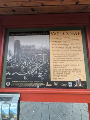 A red-framed notice board in the Peace Arch park, headed Peace Arch Centennial Commemoration, welcoming visitors in several languages.