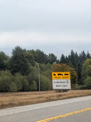 A yellow and white roadside sign reading TRUCKS - BUSES, To USA Border, NEXT EXIT, with truck and bus symbols and a highway 15 shield, standing in dry grass before a wall of trees under grey cloud.