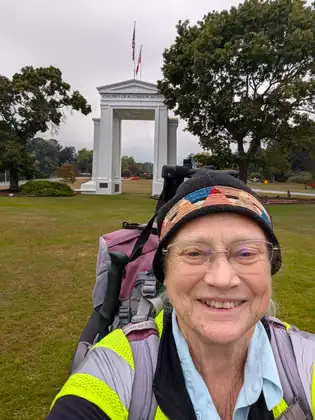 Jeana in a knitted hat and high-visibility vest, smiling with her pack on and the Peace Arch behind her on September 23, 2026.