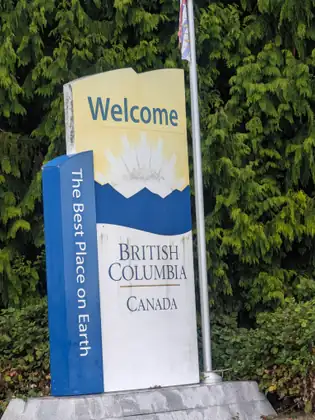 A tall sign reading Welcome, British Columbia, Canada, with The Best Place on Earth running down its blue edge, against a green cedar hedge.