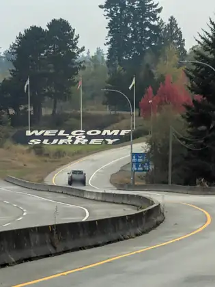 A curving divided highway with a pickup truck heading away along it, toward big white letters on a black band across a grassy slope reading WELCOME TO SURREY, with flags, evergreens and a red-leaved tree behind.