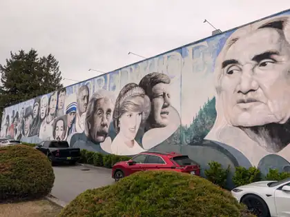 A mural the length of a building, painted with the faces of famous people from around the world under the word WORLDWIDE, parked cars and rounded hedges in front of it under a grey sky.