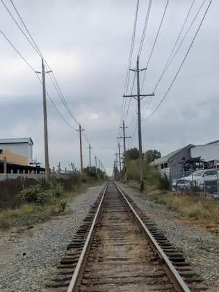 A single railway track running straight ahead between gravel shoulders and wooden power poles, a warehouse on one side and a fenced yard of parked trucks on the other.