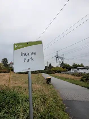 A City of Surrey sign reading Inouye Park beside a paved path, power lines and a steel transmission tower beyond the grass.