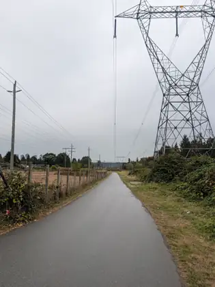 A paved path running straight ahead beneath a huge steel transmission tower, a wire fence and power poles along one side and apartment towers on the far horizon.