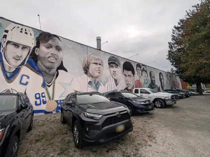 Further along the same mural, a row of painted portraits of sports figures, among them a hockey captain wearing number 99, an athlete with a gold medal and a baseball player in a New York cap, above a gravel lot of parked cars and a tree turning red.