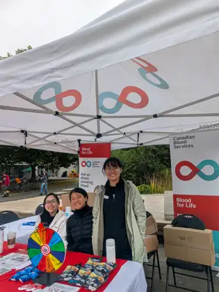 Three smiling young women at a Canadian Blood Services table under a white tent, one standing in a rain jacket, beside a prize wheel and a Blood for Life banner reading Ask us about donating blood.