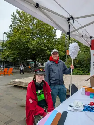 A young person in a red hoodie and a peaked cap sitting at the tent table, and behind them a smiling man in a grey cap and red scarf holding a giant cotton swab on a pole.