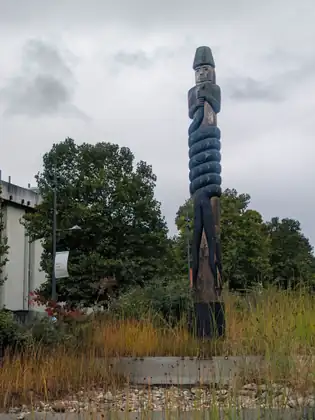 A tall carved wooden pole on the campus, a figure in a tall hat at the top grasping a long carved form that coils round and round the pole below, standing in golden grasses under a grey sky.