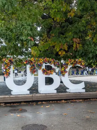 Big white letters spelling UBC, garlanded with sunflowers and autumn flowers, under a spreading oak, with students walking across the plaza behind.