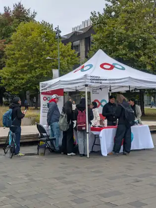 The Canadian Blood Services tent on a paved plaza, people with backpacks gathered at its table and one more standing by with a bicycle, looking at a phone.