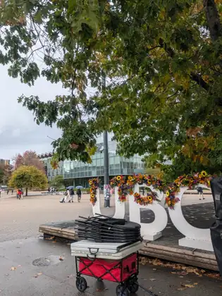 Folding chairs and a table packed up on a red pull wagon beside the flower-trimmed UBC letters, a glass-fronted building and people with umbrellas beyond.