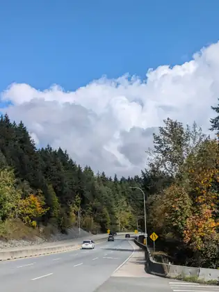 A highway sweeping round a bend between wooded hills, a towering white cloud above it and a few cars on the road.