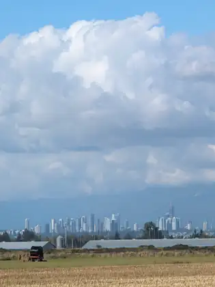 The towers of a distant city skyline across flat farmland, long low metal-roofed barns and a silo in front of it, under a mass of white cloud, a red irrigation hose reel on the grass beyond a stubble field.