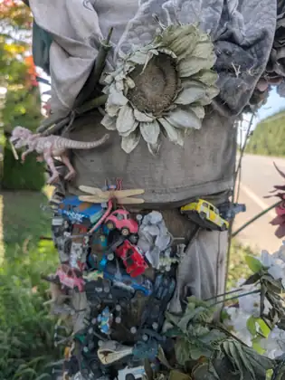 A roadside post covered in toy cars nailed on one above another, with a plastic dinosaur, a wooden dragonfly and faded fabric flowers, the road running past behind it.