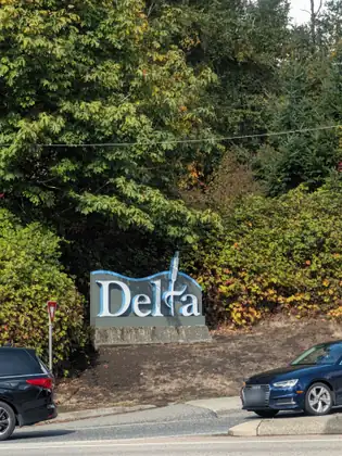 The Delta city sign, white letters edged in blue on a grey panel set on a stone base, in front of big leafy trees and shrubs turning orange, with cars on the road below it.