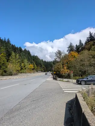 A wide highway running between wooded hills touched with yellow and orange, a dark SUV at a stop sign beside a crosswalk on a side road, and a long white cloud over the trees.