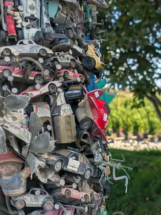 A close view of a roadside post packed with dusty toy cars held on by big nails, with a red toy airplane, a toy police car, a frayed fabric flower and a metal insect among them, a tree and potted shrubs blurred behind.