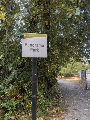 A City of Surrey sign reading Panorama Park at the start of a gravel path under leafy trees, a chain-link fence running alongside.
