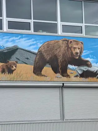 A mural painted along the side of a building, a big brown bear with one paw on a grey rock and a cub following it through golden grass, mountains and a blue sky behind them, windows above and metal shutters below.