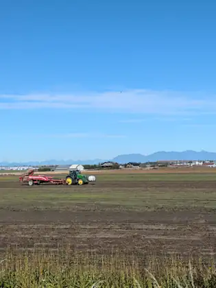 A green tractor pulling a red harvester across a bare field, farm buildings and a blue mountain range beyond under a clear sky.