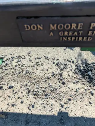 The edge of a wooden bench on the trail with a brass-lettered plaque, partly out of the frame, beginning with the name Don Moore, above sunlit gravel.