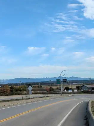 A road curving past a concrete divider toward a junction, with flat fields and a long blue mountain range beyond under a wide, wispy sky.