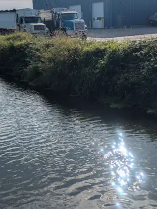 Morning sun glittering on a narrow channel of water below a grassy bank, two big trucks parked outside a warehouse above it.