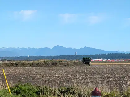 Flat harvested fields stretching to a blue wall of mountains, a green farm machine working in the distance and a long line of red shipping containers on a train beyond it.