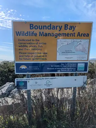 A yellow sign beside the trail reading Boundary Bay Wildlife Management Area, dedicated to the conservation of native wildlife, plants, fish and their habitats, with a map of the bay.