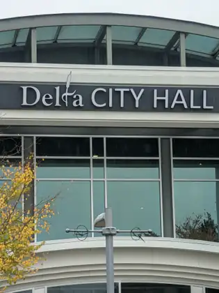 The words Delta City Hall in white letters across the front of a curved building of glass and pale concrete, a lamp post and a tree with yellow leaves in front of it.