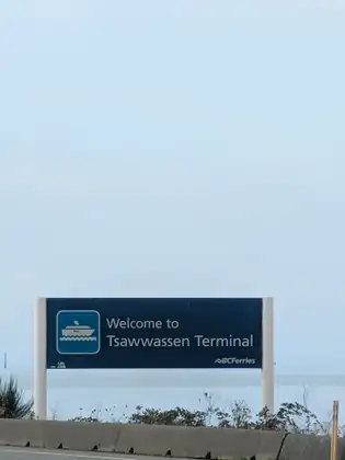 A dark blue BC Ferries sign with a white ferry symbol reading Welcome to Tsawwassen Terminal, standing above a concrete barrier and roadside weeds against pale sky and flat, misty water.