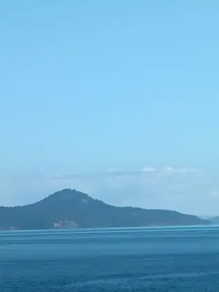A forested hill rising to a rounded peak above a long wooded shoreline across deep blue water, a small white boat near the shore, under a hazy pale blue sky with a few small white clouds.