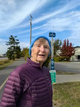 An older woman in a blue knitted hat and a plum quilted jacket stands beside a paved trail under streaky cloud, with signs reading Mills Road, 25 km and Sidney behind her and a red-leaved tree beyond.