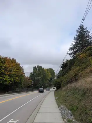 A two-lane road climbing past autumn trees and a grassy bank, cars heading away along it and a sidewalk on the right.