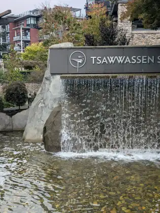 A sign beginning with the word Tsawwassen above a water feature spilling over a stone wall into a pebbled pool, multi-storey buildings with balconies behind.