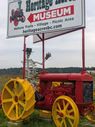 A skeleton in a black wide-brimmed hat waving from the seat of an old red Fordson tractor with yellow spoked wheels, beneath a sign reading Heritage Acres, MUSEUM, Nature Trails - Village - Picnic Area and heritageacresbc.com.