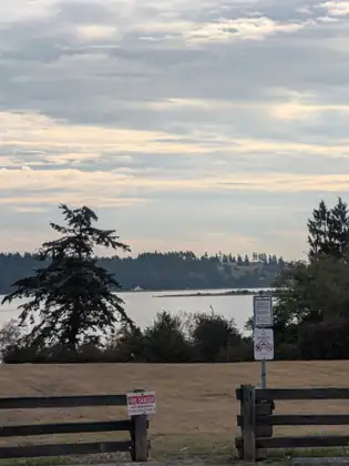 Through a gap in a split-rail fence, a dry lawn stretches to dark shrubs and calm water with a low wooded shore beyond, a tall ragged evergreen at left and signs reading FIRE DANGER and NO SMOKING.