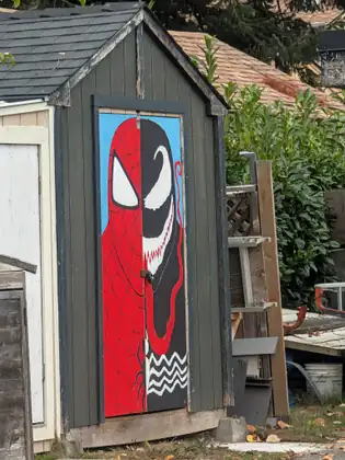 A dark grey garden shed whose door is painted with one face split down the middle, half Spider-Man’s red webbed mask and half Venom’s black face with a white eye, jagged teeth and a long red tongue, boards and lattice leaning beside it.