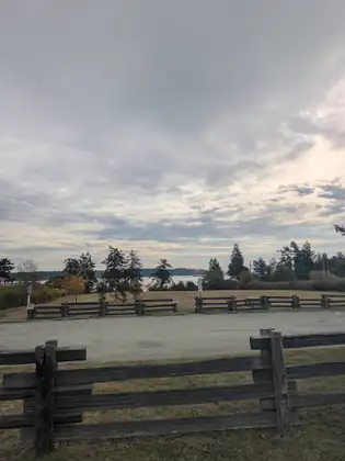 A wide gravel lot lies between two weathered split-rail fences, with a dry lawn, tall evergreens and a glimpse of calm water and a wooded shore beyond under a broad sky of gray and pale gold cloud.